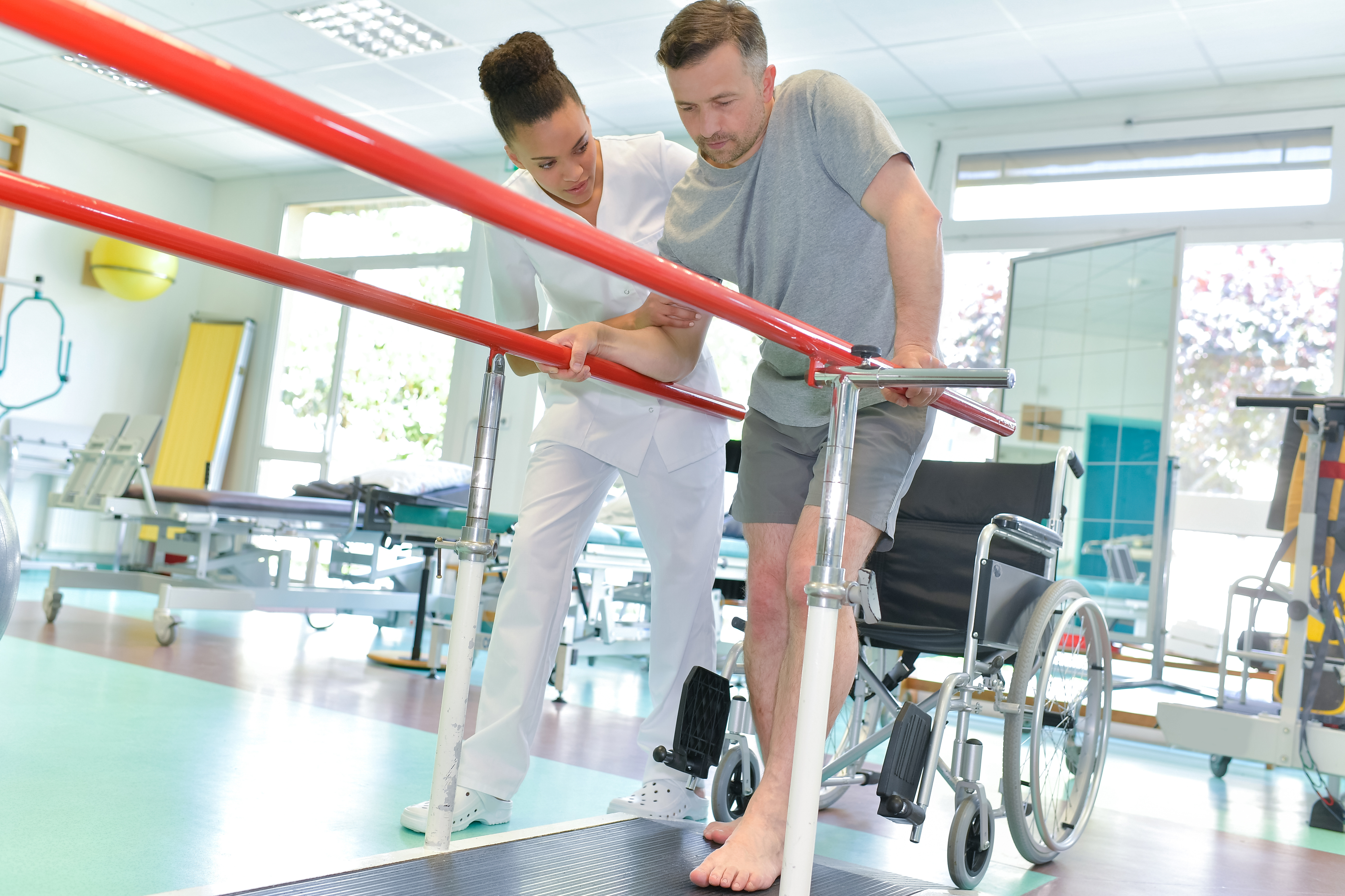 Therapist guides an adult male patient on parallel bars during a gait therapy session in a rehab gym.