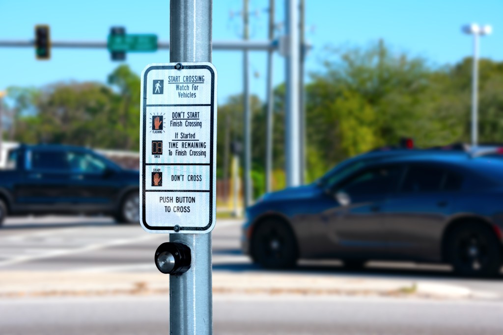 Traffic intersection pedestrian crosswalk crossing sign with signal descriptions above the request button with cars and lights in the background - Travis Legal Offices | Colorado Personal Injury Law Firm Close-up of a pedestrian crossing sign on a metal pole: ‘Start Crossing’ and ‘Push Button to Cross’ with vehicle traffic in background.