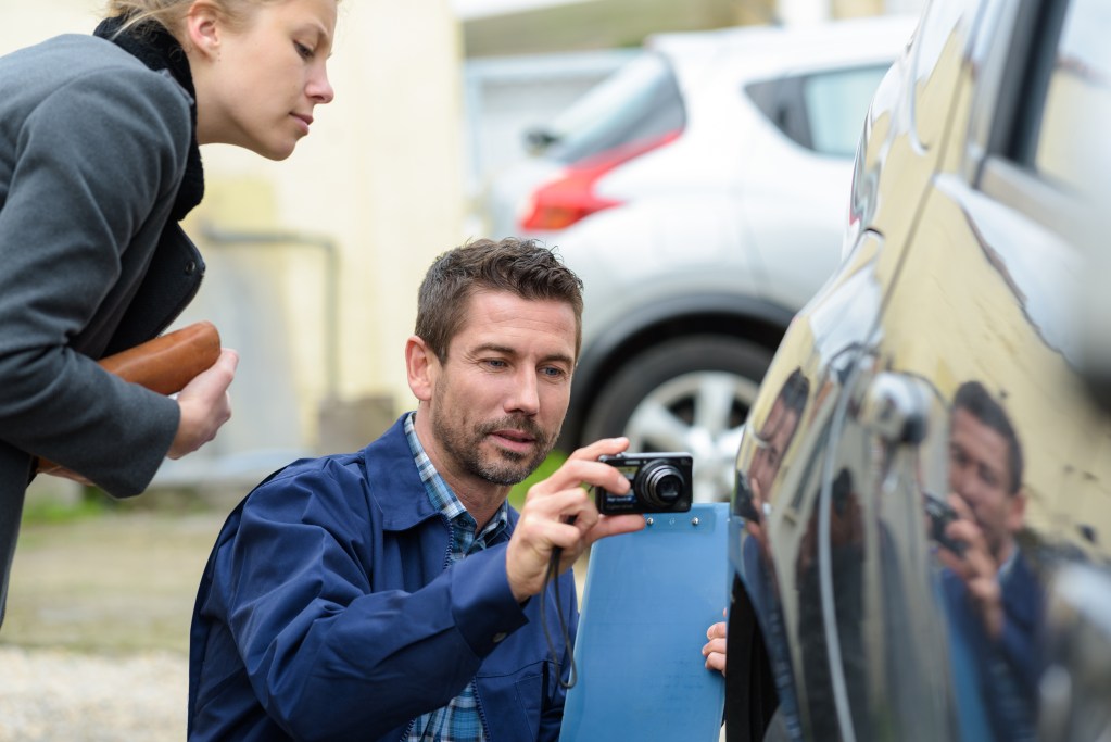 Man in a blue jacket uses a camera to photograph a blue car door while a woman in a gray coat watches nearby.