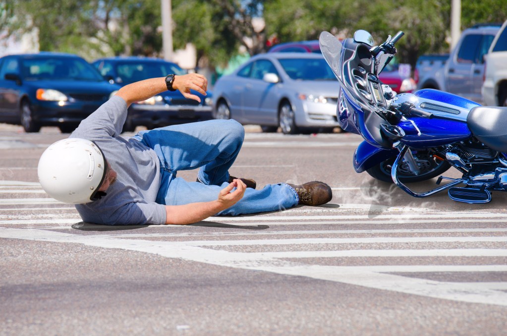 Motorcycle Wreck in Busy Intersection - Travis Legal Offices | Colorado Personal Injury Law Firm Man lying on his side after a motorcycle crash in a parking lot, helmet on, next to a blue motorcycle with chrome detailing.