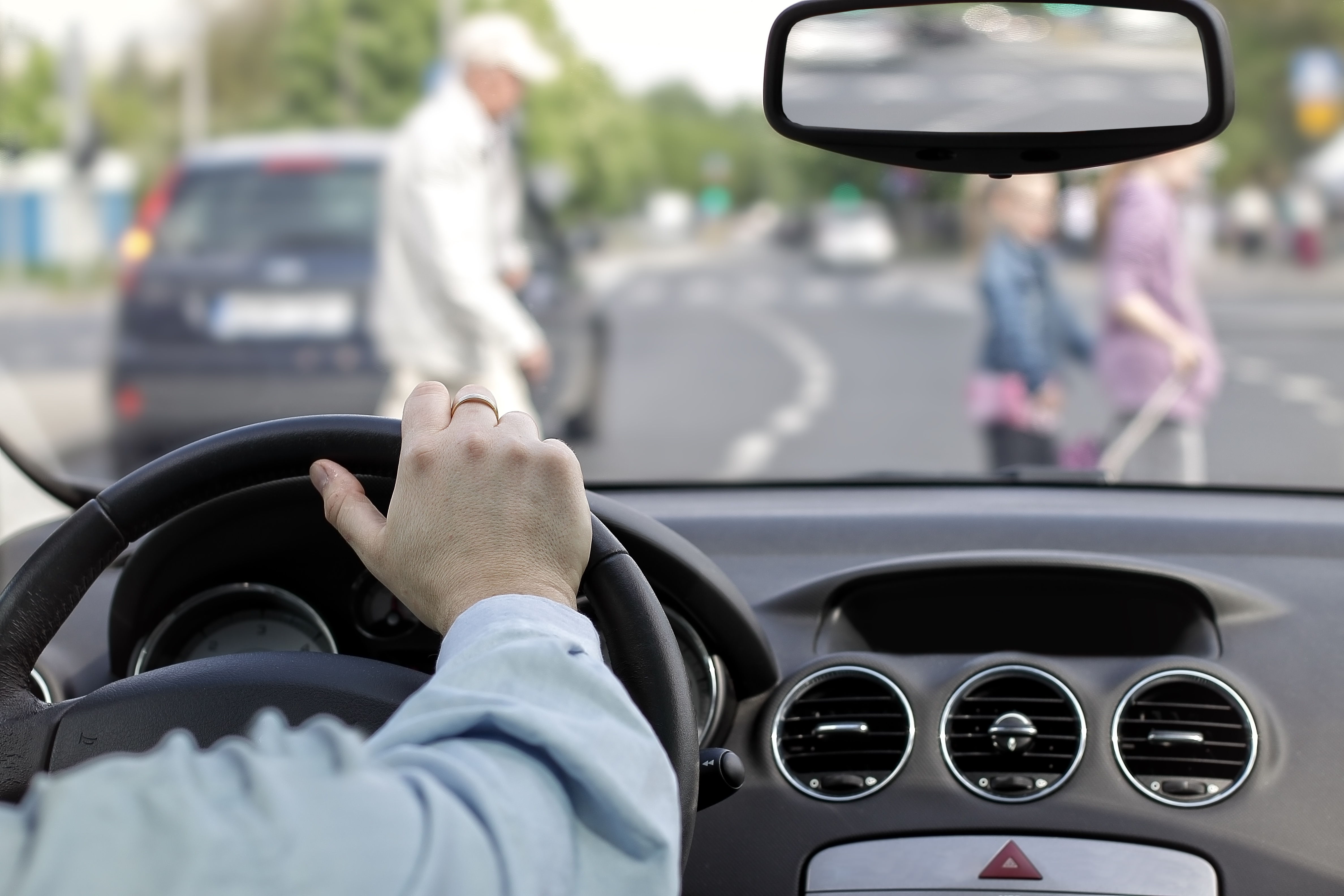 Driver's hand on steering wheel with a wedding ring, dashboard and rearview mirror showing a blurred urban street scene ahead