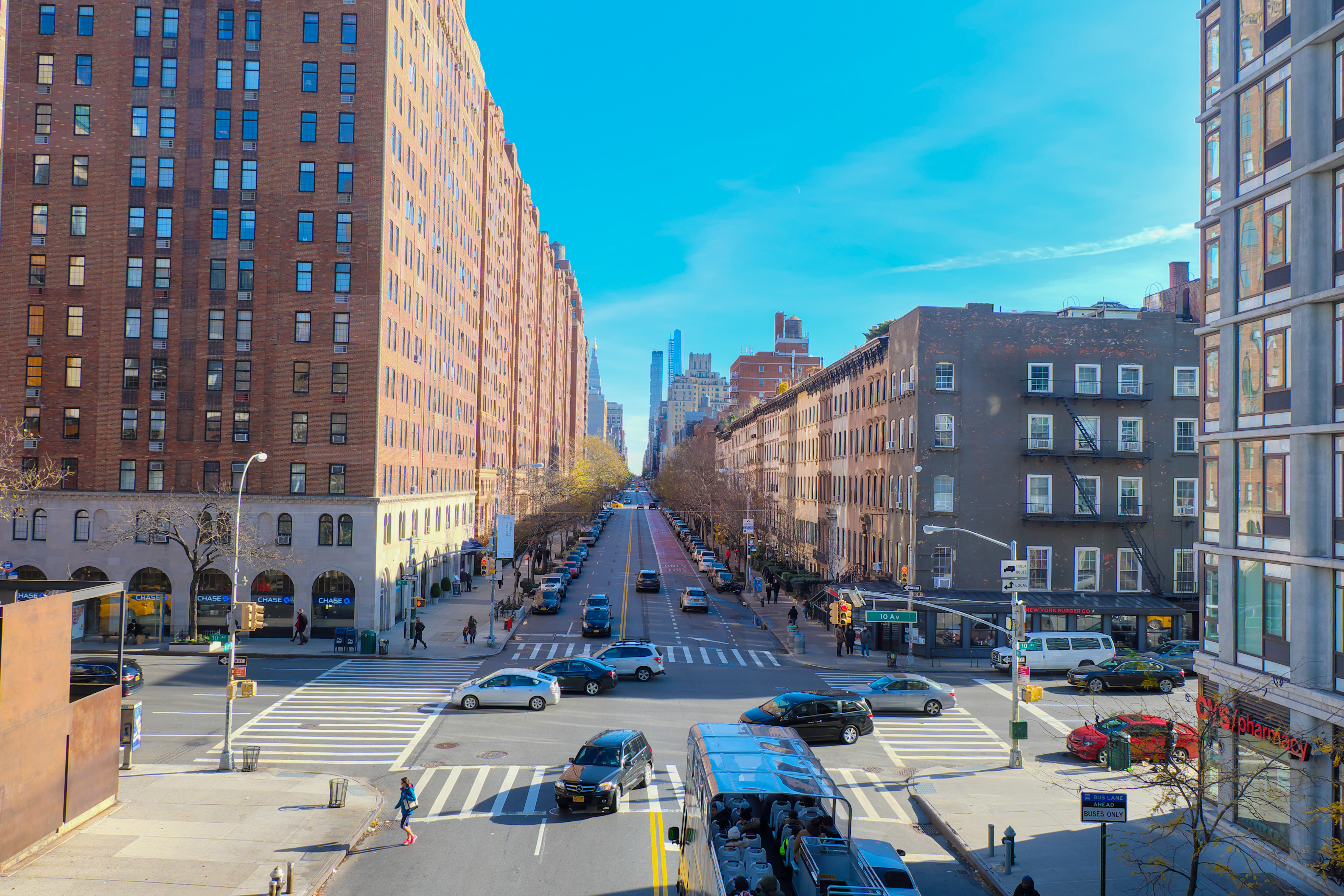 Wide city street lined with tall brick apartment buildings on both sides under a bright blue sky, with cars and pedestrians