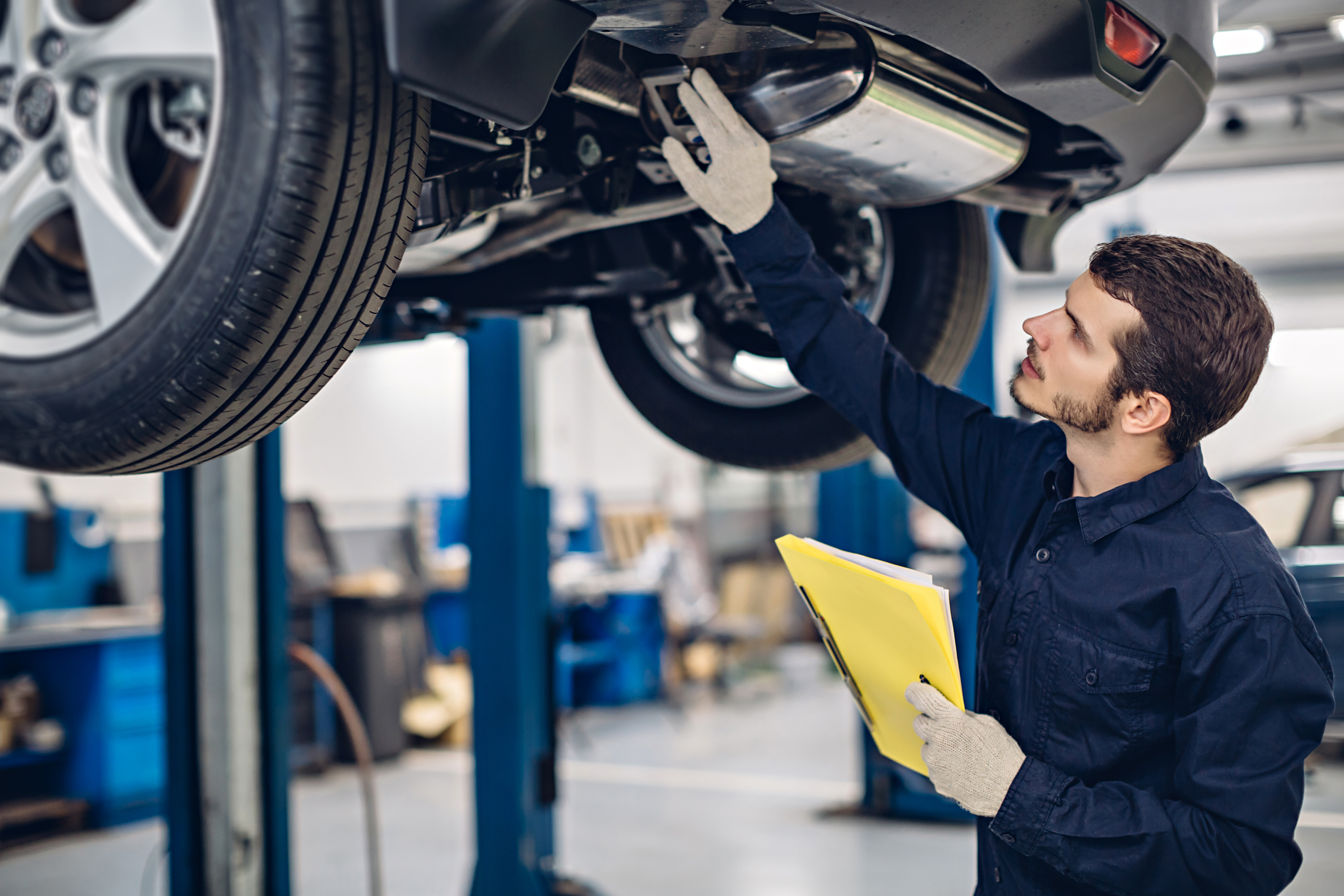 Mechanic in navy coveralls inspecting a car lifted off the ground, holding a yellow folder and wearing gloves in a workshop
